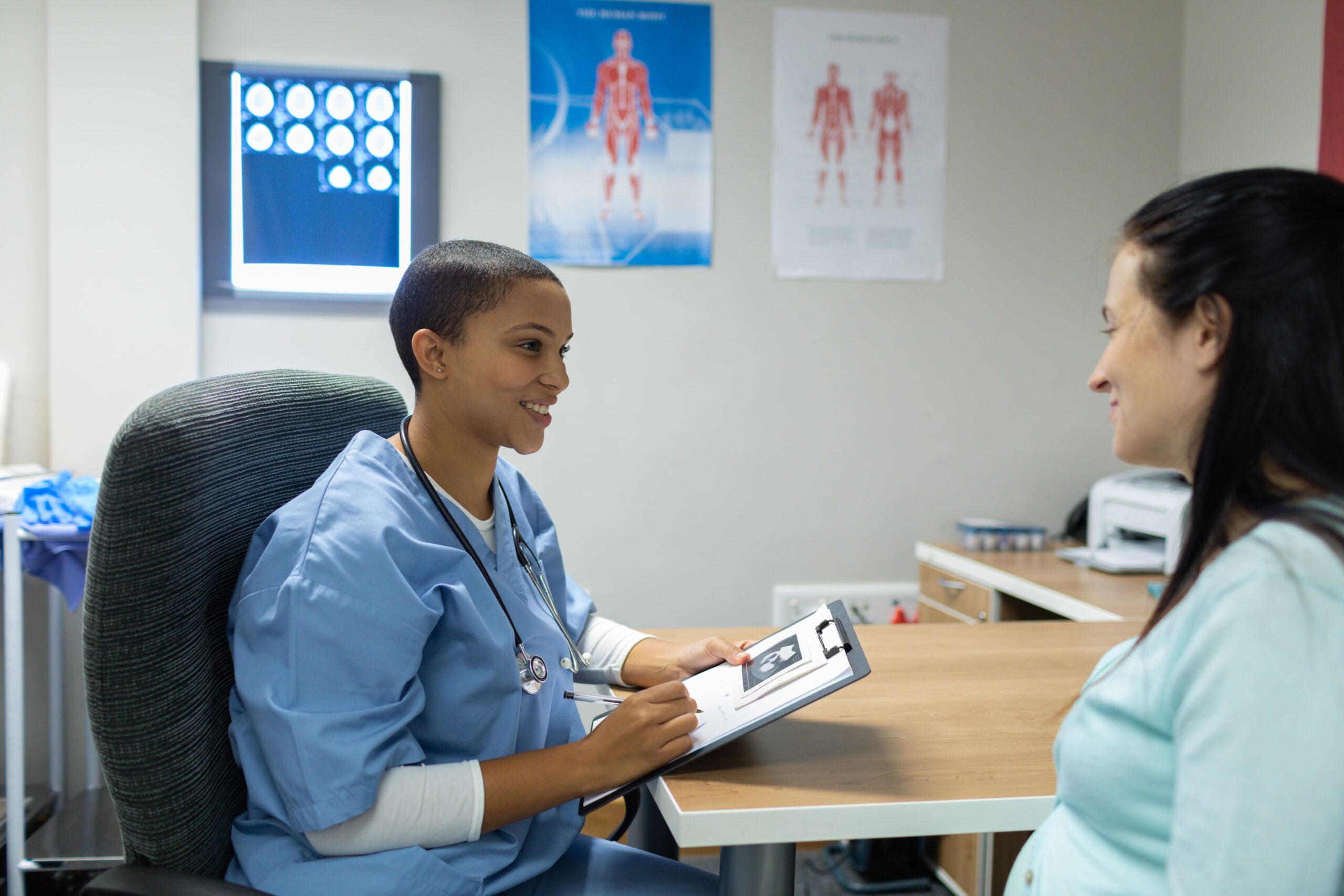 Mixed-race female doctor giving a prescription to a pregnant woman at the desk of an examination room