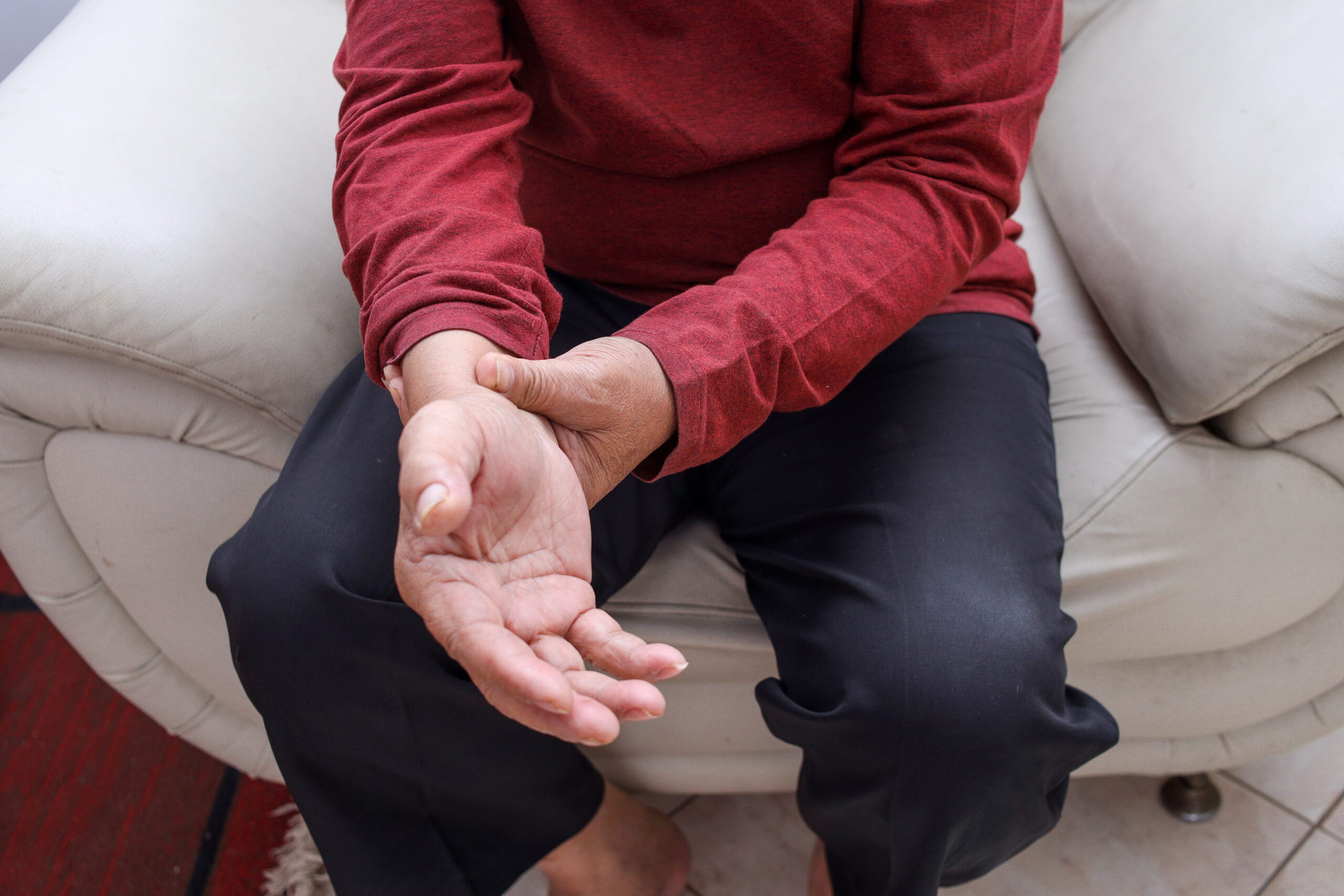 An elderly man sits on a sofa in his living room, holding and massaging his wrist, suffering from wrist joint pain.