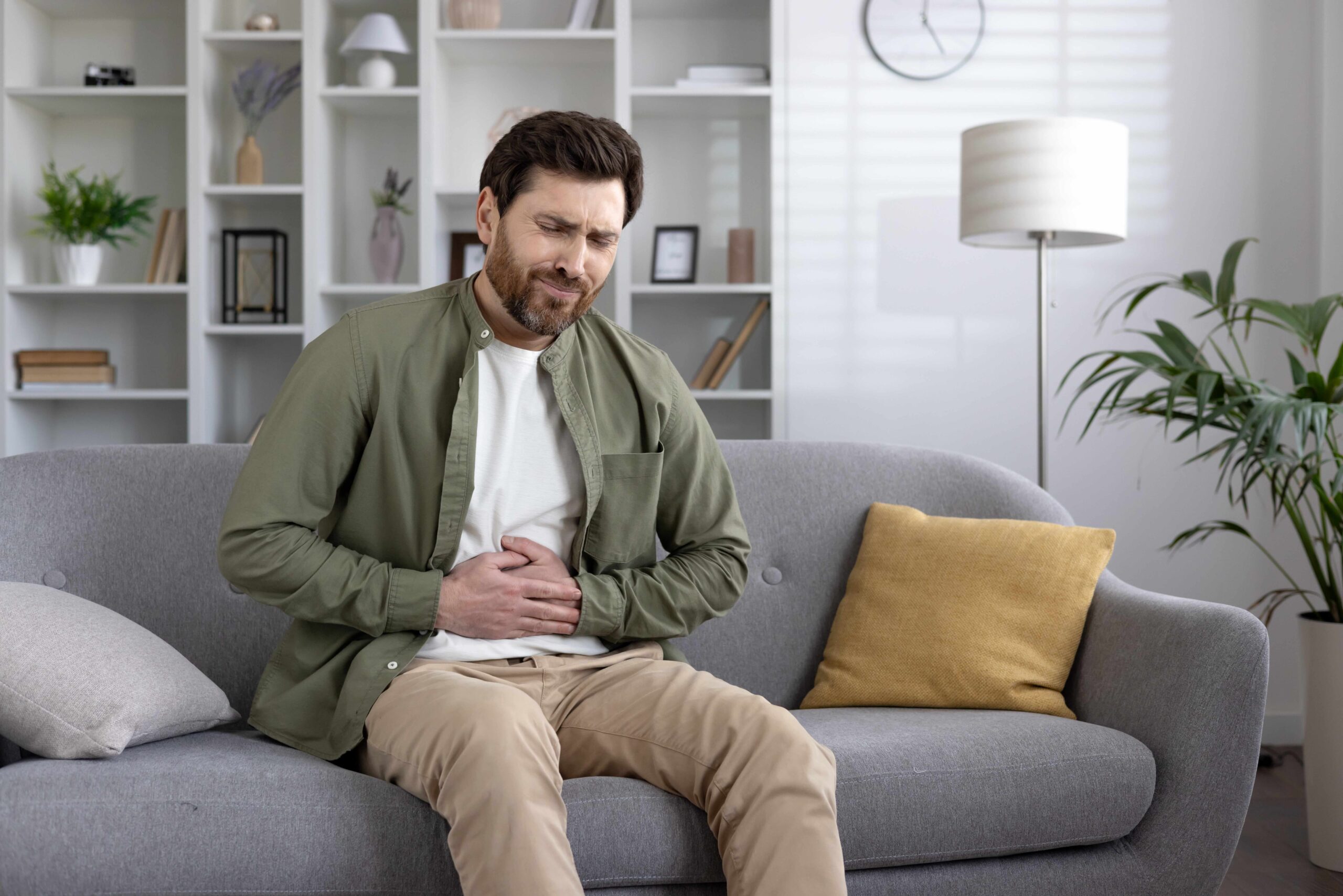 A man sitting on a couch, holding his stomach in pain at home.