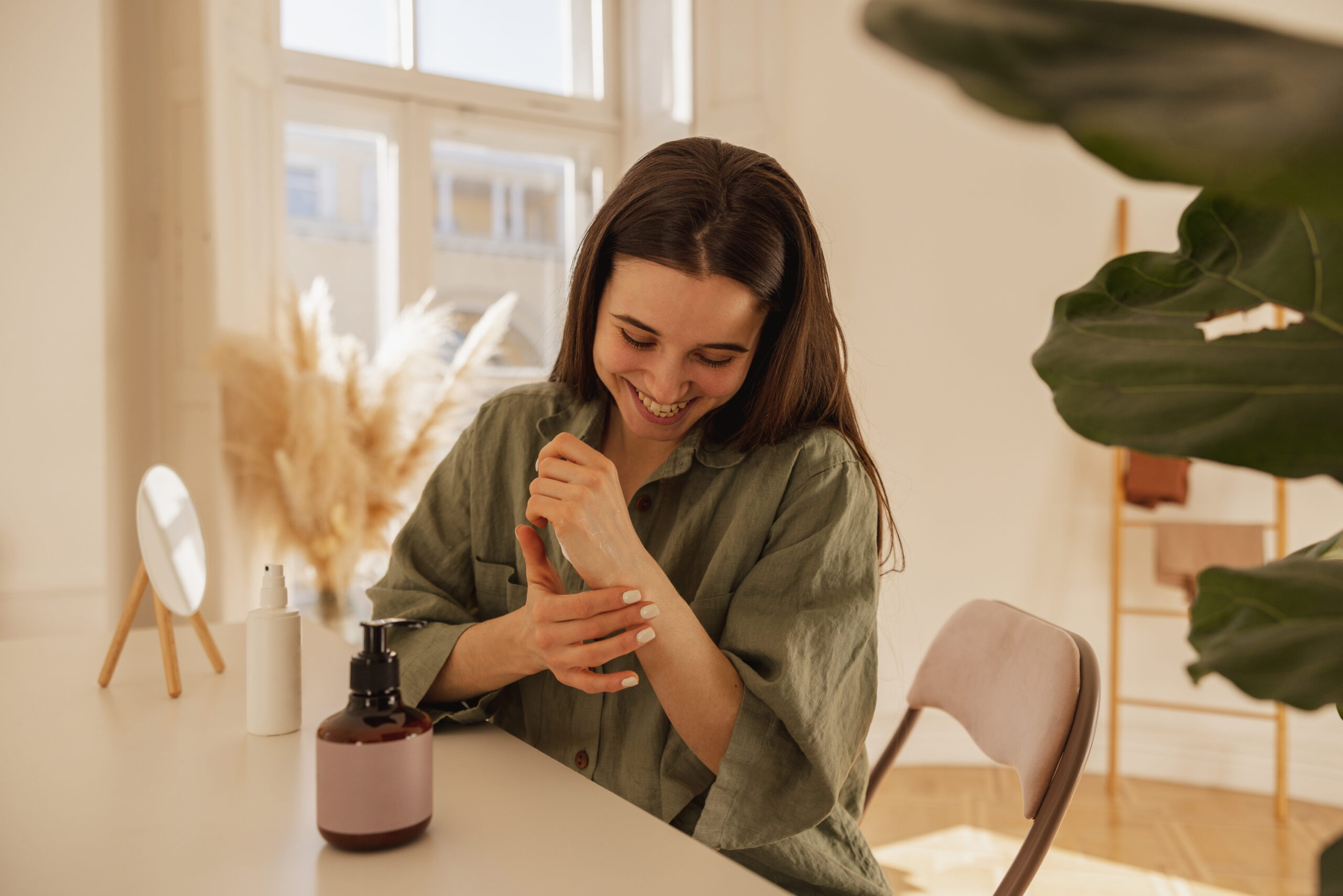 cheerful-young-caucasian-woman-applies-cream-her-hands-while-sitting-table-light-room-brunette-girl-with-smooth-skin-wears-shirt-wellness-self-care-concept